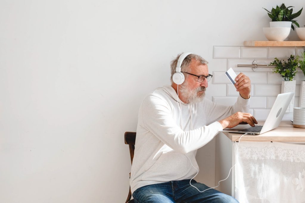 Äldre man med grått skägg och glasögon sitter vid ett bord med en bärbar dator, bär vita hörlurar och håller ett kreditkort i handen, verkar göra ett onlineköp i ett ljust kök med vita kakelväggar och växter i bakgrunden.