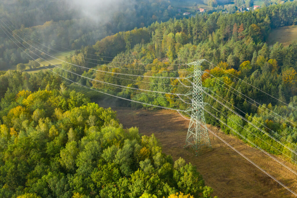 Aerial view of the high voltage power lines and high voltage electric transmission on the terrain surrounded by trees at sunlight Aerial view of the high voltage power lines and high voltage electric transmission on the terrain surrounded by trees at sunlight.