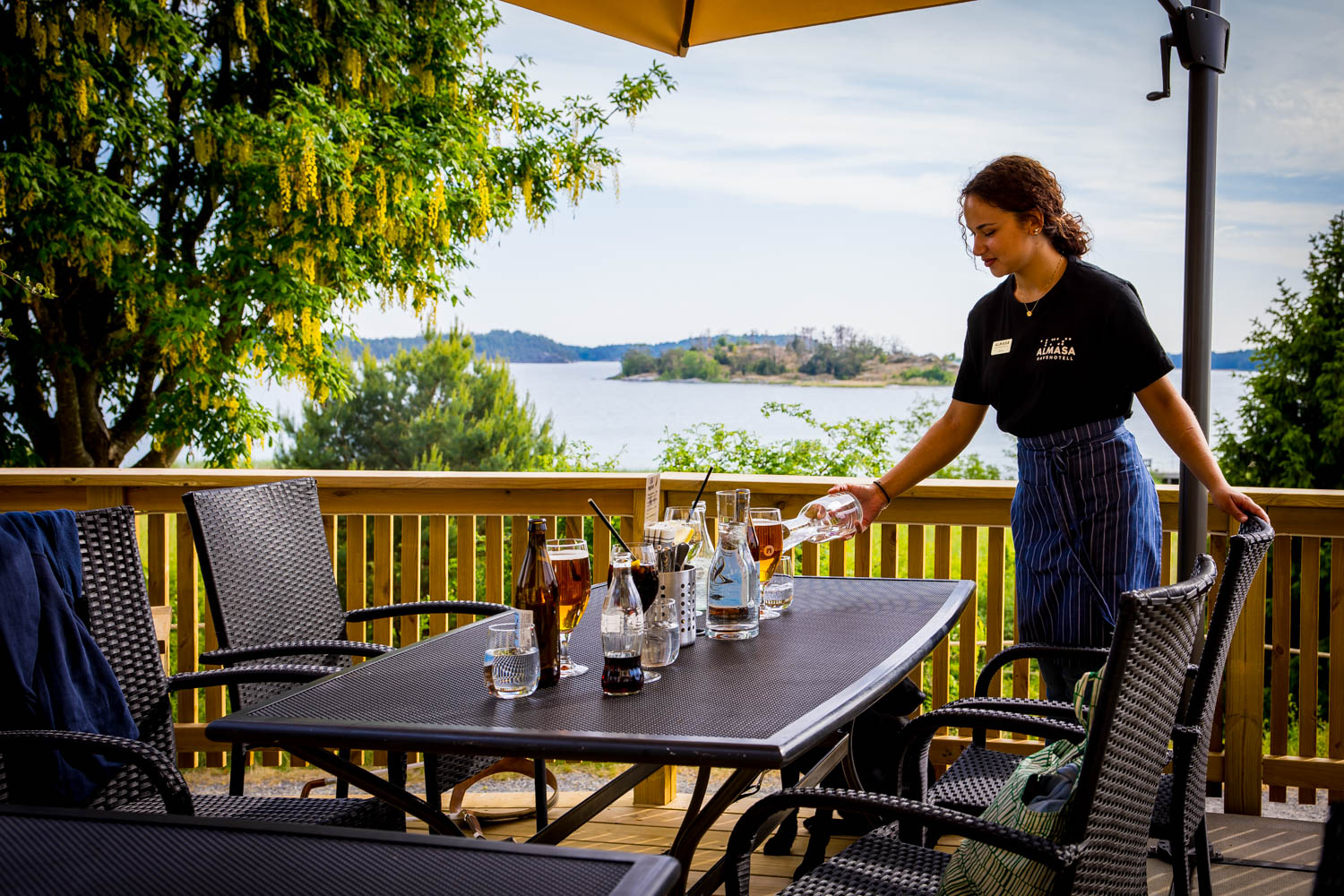 En servitris häller vatten i ett glas vid ett dukat utomhusbord på en terrass med utsikt över vatten och grönskande öar i bakgrunden.