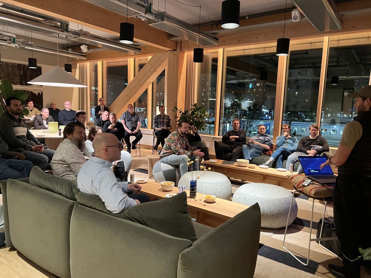 En grupp människor sitter samlade i ett öppet kontorsutrymme och lyssnar på en presentation. Föreläsaren står till höger med en laptop, och åhörarna sitter i soffor och stolar framför stora fönster som vetter mot en stadsmiljö på kvällen